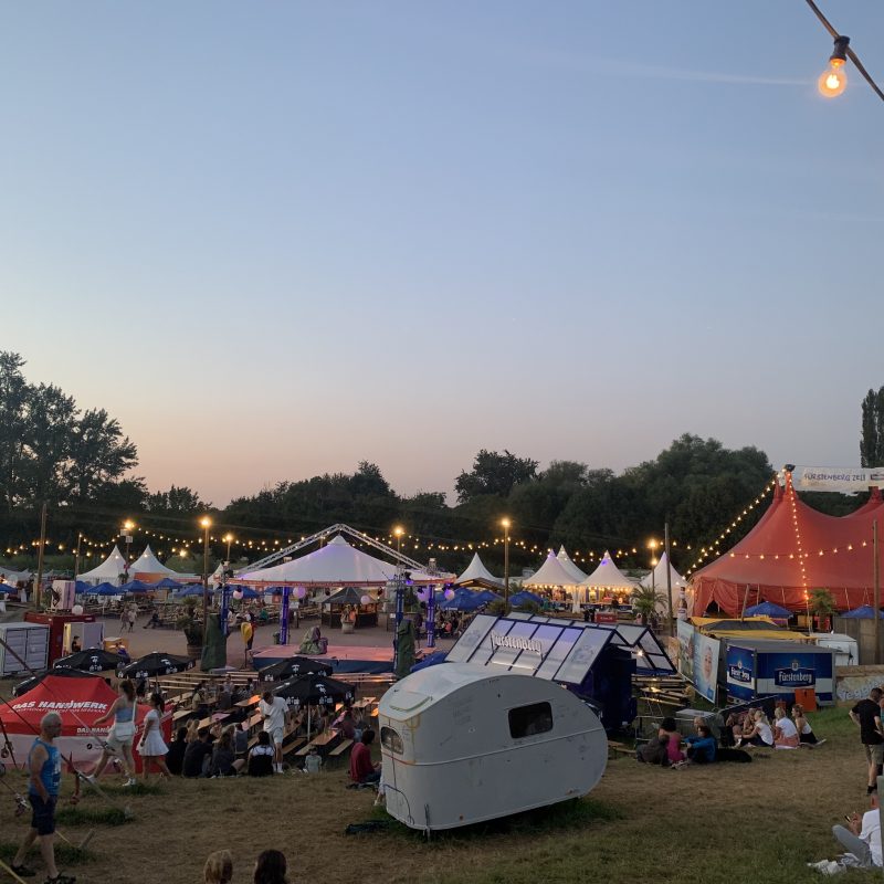 Zu sehen ist das Gelände des Zelt-Musik-Festivals. Die Zeltspitzen sind mit Lichterketten geschmückt und Menschen sitzen über das Gelände verteilt zusammen.