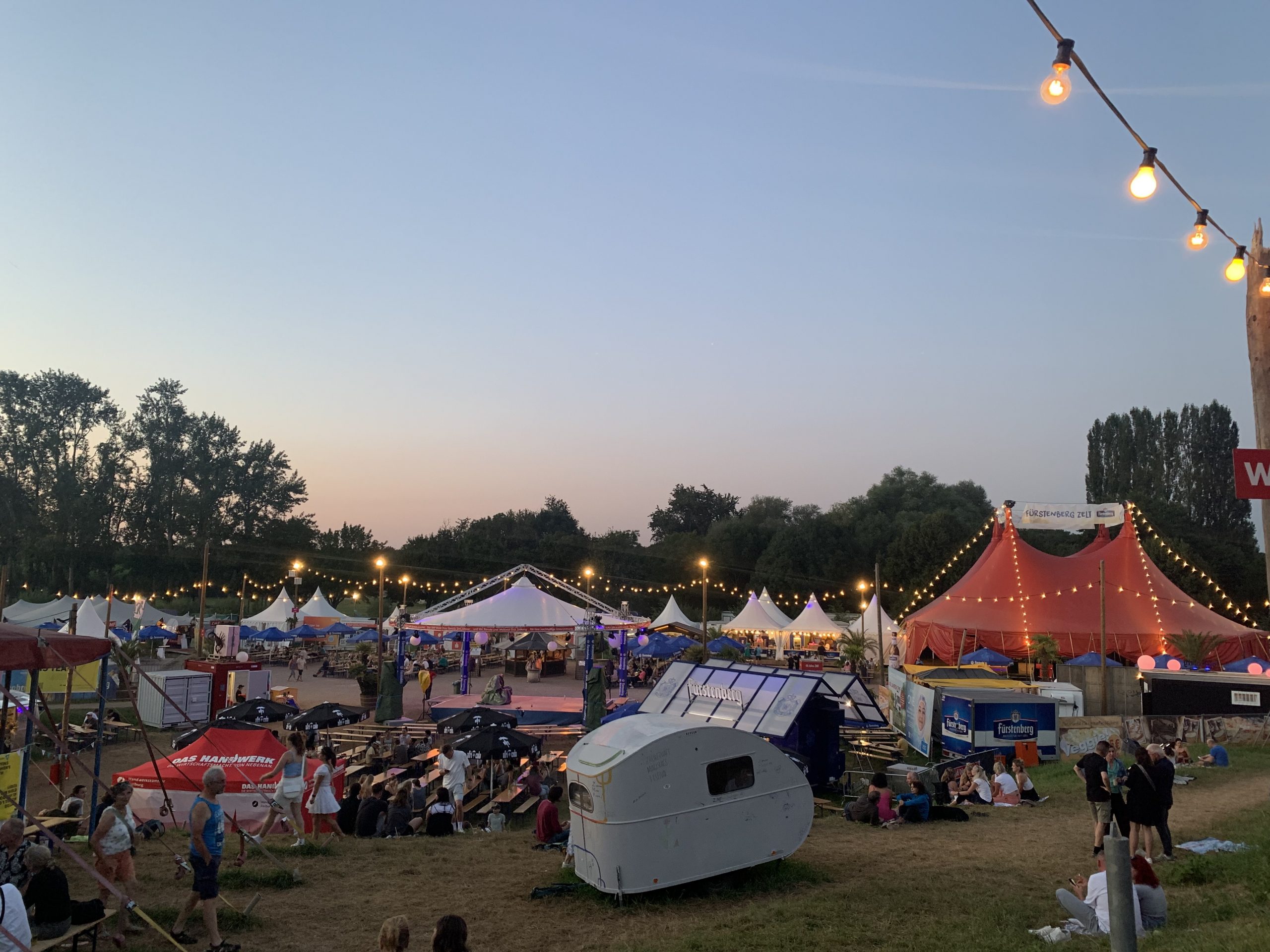Zu sehen ist das Gelände des Zelt-Musik-Festivals. Die Zeltspitzen sind mit Lichterketten geschmückt und Menschen sitzen über das Gelände verteilt zusammen.