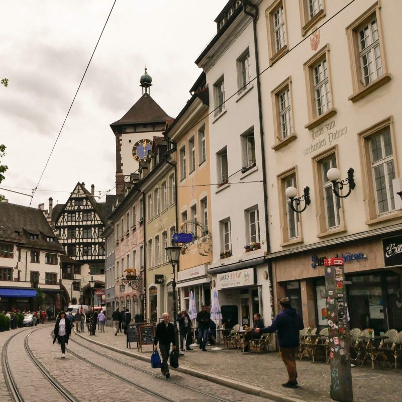 Zu sehen ist der Stadtteil Oberlinden. Die Straßenbahnschienen deuten den Weg Richtung des Schwabentores, das halb verdeckt zu sehen ist. Es ist ein geschäftiger Tag und einige Menschen bummeln durch die Straßen.