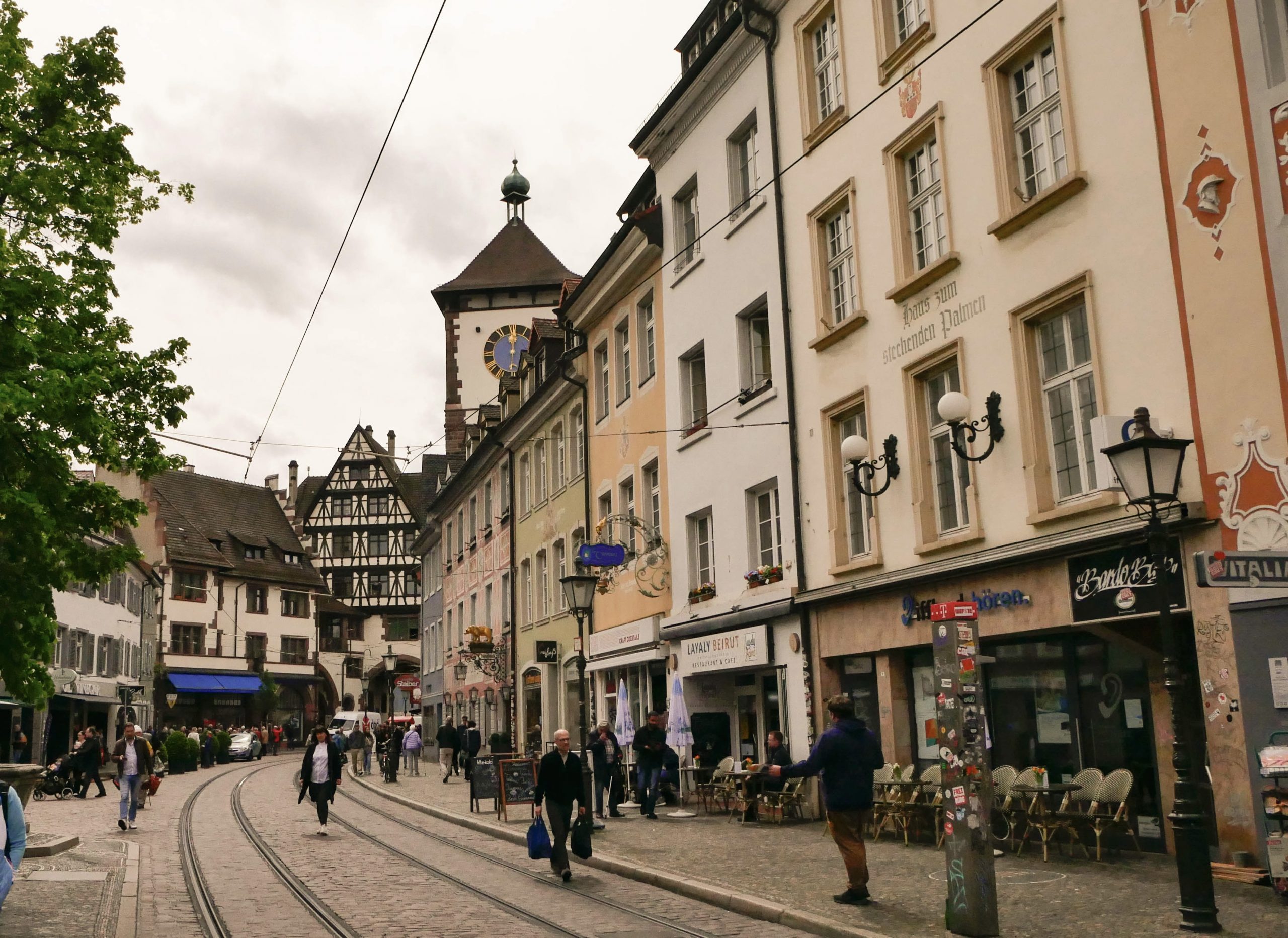 Zu sehen ist der Stadtteil Oberlinden. Die Straßenbahnschienen deuten den Weg Richtung des Schwabentores, das halb verdeckt zu sehen ist. Es ist ein geschäftiger Tag und einige Menschen bummeln durch die Straßen.