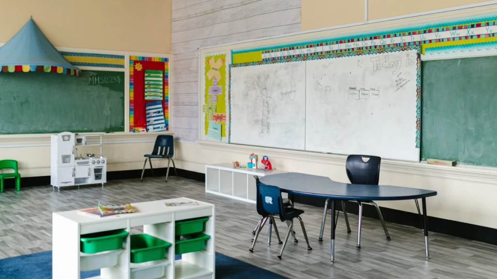 Zu sehen ist ein leeres Klassenzimmer. An den Wänden sind Tafel und Whiteboards angebracht. Es stehen kleine weiße Regal an den Wänden und eines mitten im Raum. Es steht außerdem ein Tisch mit zwei Stühlen im Raum und ein weiterer Stuhl in der Ecke.