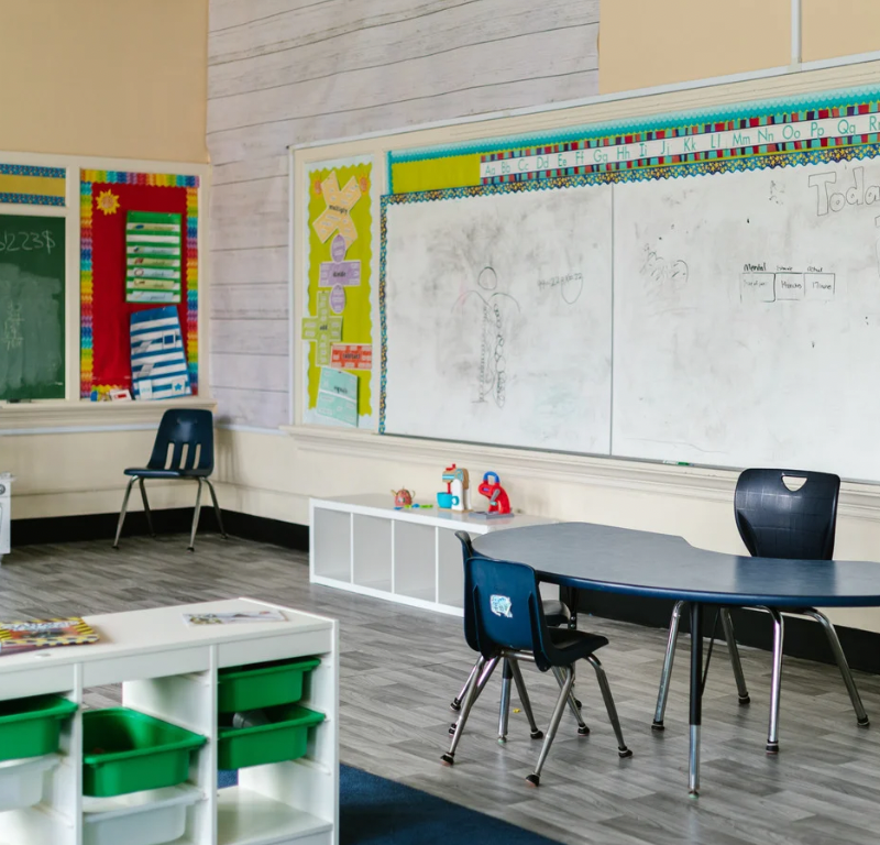Zu sehen ist ein leeres Klassenzimmer. An den Wänden sind Tafel und Whiteboards angebracht. Es stehen kleine weiße Regal an den Wänden und eines mitten im Raum. Es steht außerdem ein Tisch mit zwei Stühlen im Raum und ein weiterer Stuhl in der Ecke.